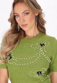 Zielony sweter z dzianiny, ozdobiony perłami, z czarnymi akcentami w postaci kokardek oraz teksturowanym wykończeniem. Krótkie rękawy i okrągły dekolt.