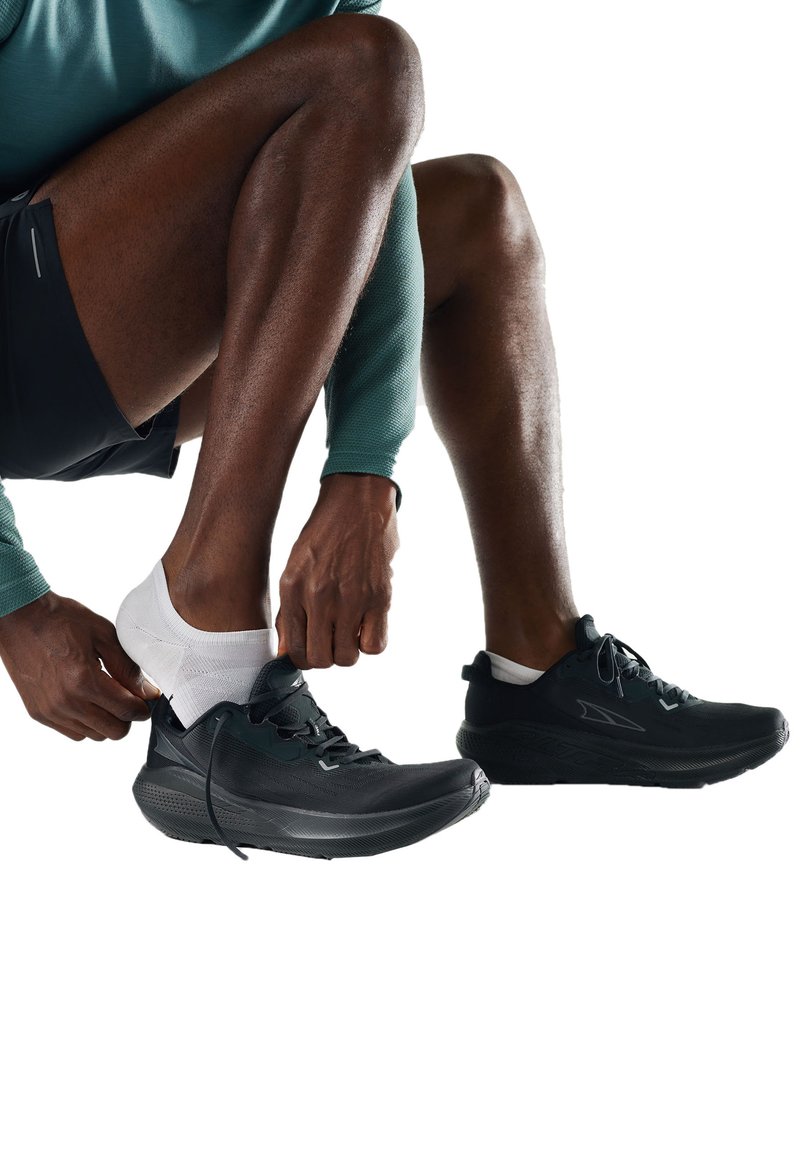 Osoba nosząca białe skarpetki zakłada czarne buty do biegania, ubrana w czarne szorty i turkusową koszulkę z długim rękawem, siedząc na tle białego tła.