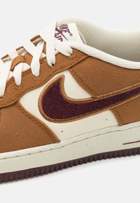 Tenis de lona castanhos com detalhes em creme, design de swoosh vinho, superfície texturizada, sola de borracha e atacadores contrastantes.