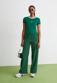 Zelené tričko s logom "Nike", skombinované so voľnými zelenými nohavicami, bielymi teniskami a slnečnými okuliarmi. Model drží malú striebornú tašku.