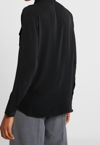 Persona vistiendo una blusa negra de manga larga con puños abotonados y pantalones grises texturizados, mostrada de espaldas contra un fondo liso.