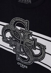 Juodas organinės medvilnės marškinėliai su dideliu „GUESS“ logotipu, puoštu rhinestones, ir baltais juostelių akcentais tamsiame fone.