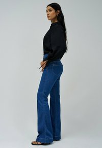 Blusa negra de manga larga con textura y puños fruncidos, combinada con jeans de campana azules de cintura alta. La modelo lleva sandalias negras con tiras.