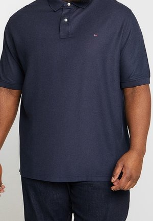 Mand iført en mørkeblå kortærmet polo med to knapper og mørke bukser, stående foran en ensfarvet baggrund.