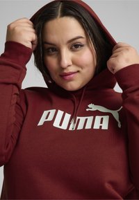 Felpa con cappuccio color bordeaux realizzata in morbido tessuto, caratterizzata da un grande logo bianco "PUMA" e da una silhouette di gatto. Cappuccio tirato su intorno al volto.
