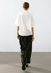 Camicia bianca a maniche corte, con bottoni, realizzata in tessuto leggero, abbinata a pantaloni neri larghi e tacchi neri spessi.