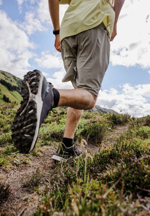 Zwarte en grijze trailrunningschoenen met gestructureerde zolen, gedragen met grijze shorts, stapend op een paden van aarde omringd door groene vegetatie.