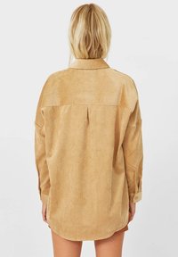 Khaki overhemd gemaakt van zachte, gestructureerde stof. Het heeft een klassieke kraag, knoopmanchetten en een afgeronde zoom met een plooidetail aan de achterkant.