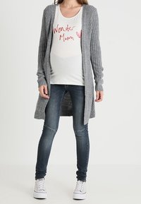 Šedý žebrovaný cardigan přes bílé tričko s nápisem "Wonder Mum", doplněný o tmavě modré skinny džíny a bílé tenisky.