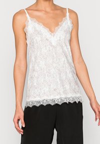 Žena v bílé camisole s krajkovým lemem, tenkými ramínky a jemným tečkovaným vzorem, spárovaná s volnými černými kalhotami, na světlém pozadí.