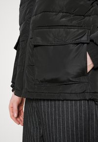 Persona che indossa una giacca nera trapuntata con tasche a pattina e pantaloni a righe, con una mano nella tasca della giacca.