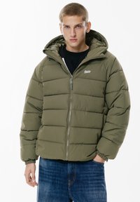 Giubbino imbottito verde oliva, con chiusura a zip, cappuccio e design trapuntato. Abbinato a jeans in denim blu.