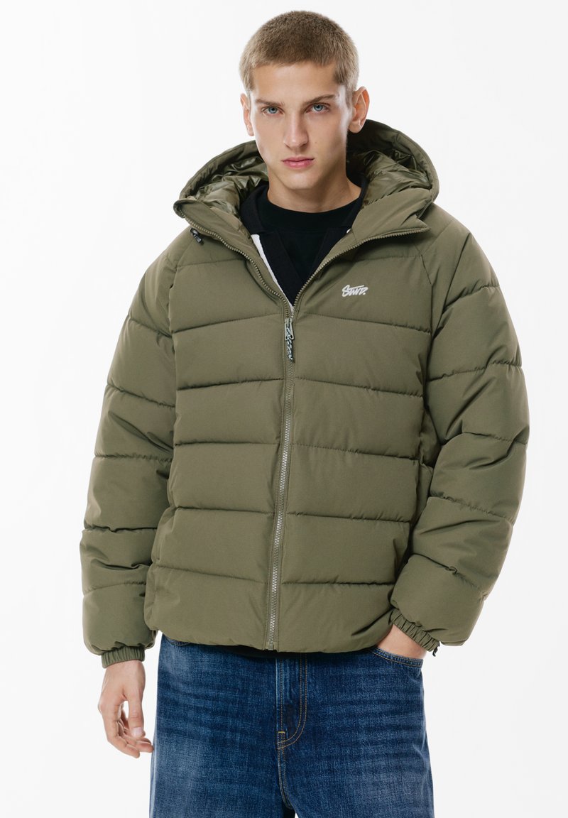 Giubbino imbottito verde oliva, con chiusura a zip, cappuccio e design trapuntato. Abbinato a jeans in denim blu.