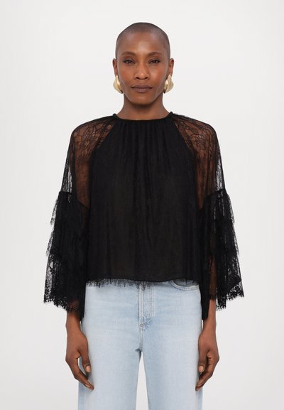 Blouse en dentelle noire avec tissu transparent, manches en évêque et accents froncés. Col haut ; léger et texturé, associé à un jean clair.