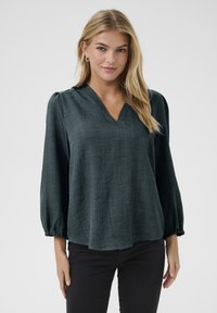 Blusa lunga a maniche con col V di colore verde scuro, con polsini arricciati; tessuto testurizzato; vestibilità comoda. Indossata con pantaloni neri.