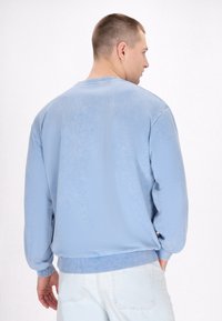 Felpa blu con collo rotondo, polsini e orlo ribattuti, e una texture morbida. La vista posteriore mostra un tessuto liscio con cuciture minime.