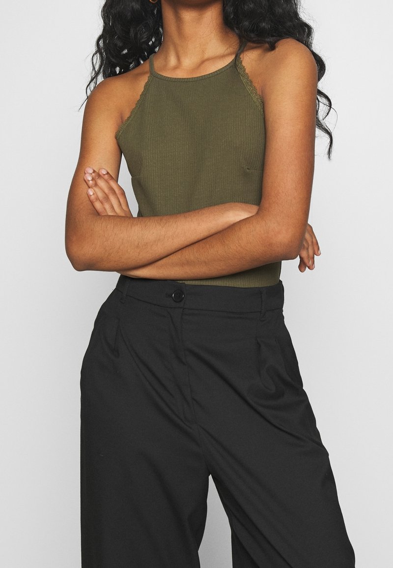 Femme aux longs cheveux bouclés portant un haut sans manches vert olive et un pantalon noir taille haute, les bras croisés sur la poitrine.