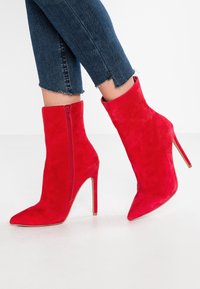 Stivaletti alla caviglia in suede rosso con punta affusolata e tacco a stiletto. Presentano una zip laterale e una texture liscia. Indossati con jeans di denim tagliati.
