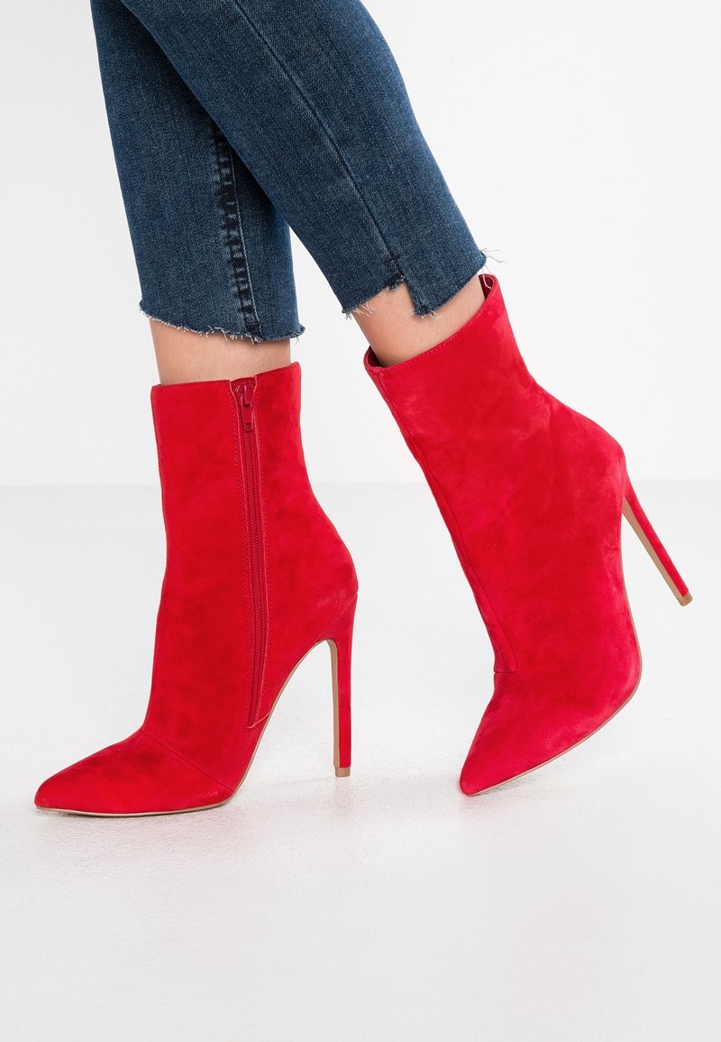 Stivaletti alla caviglia in suede rosso con punta affusolata e tacco a stiletto. Presentano una zip laterale e una texture liscia. Indossati con jeans di denim tagliati.