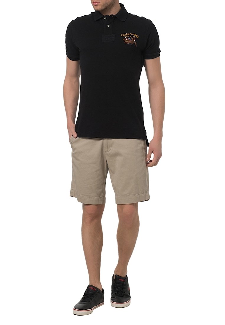Homme portant un polo noir avec le logo Ralph Lauren, un short beige à hauteur des genoux et des baskets noires, debout les mains dans les poches.