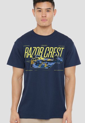 Man wearing navy blue t-shirt with yellow and blue spaceship graphic and large text "Razor Crest" on front.