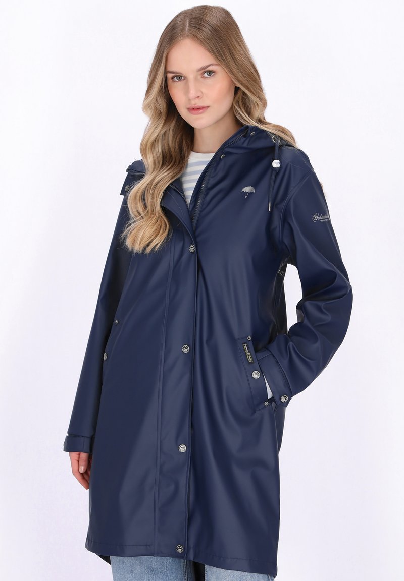 Donna con lunghi capelli biondi che indossa un impermeabile blu scuro con cappuccio, camicia a righe e jeans azzurri, in piedi con una mano in tasca.