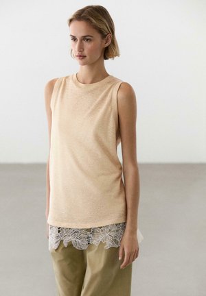 Giovane donna con top beige senza maniche e pantaloni oliva con dettaglio in pizzo bianco, in piedi su sfondo chiaro e uniforme.