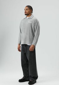 Grijze fleece trui met kraag, gestructureerd oppervlak, geborduurd logo, gecombineerd met losse zwarte jeans en zwarte veterschoenen.