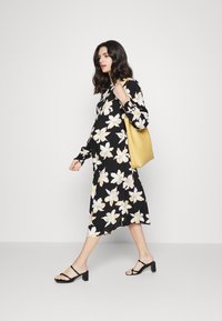Vestido preto floral com flores brancas e amarelas, mangas longas e um corte descontraído, combinado com uma bolsa amarela e sapatos de salto pretos com tiras.
