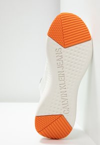 Sole of white shoe with textured grip and orange tread tips, embossed with "CALVIN KLEIN JEANS" branding.