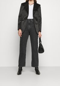 Blazer negro de satén con solapa puntiaguda, camiseta blanca ajustada, jeans de mezclilla negros de cintura alta, botas negras de tacón y un bolso pequeño negro.