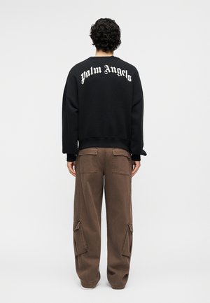 Person stående med ryggen til iført en sort sweatshirt med teksten "palm Angels" og løse brune cargobukser med sidelommer.