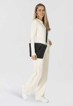 Femme avec de longs cheveux blonds portant un pantalon large crème et un pull, des baskets blanches, portant un sac à bandoulière en cuir noir, souriante.