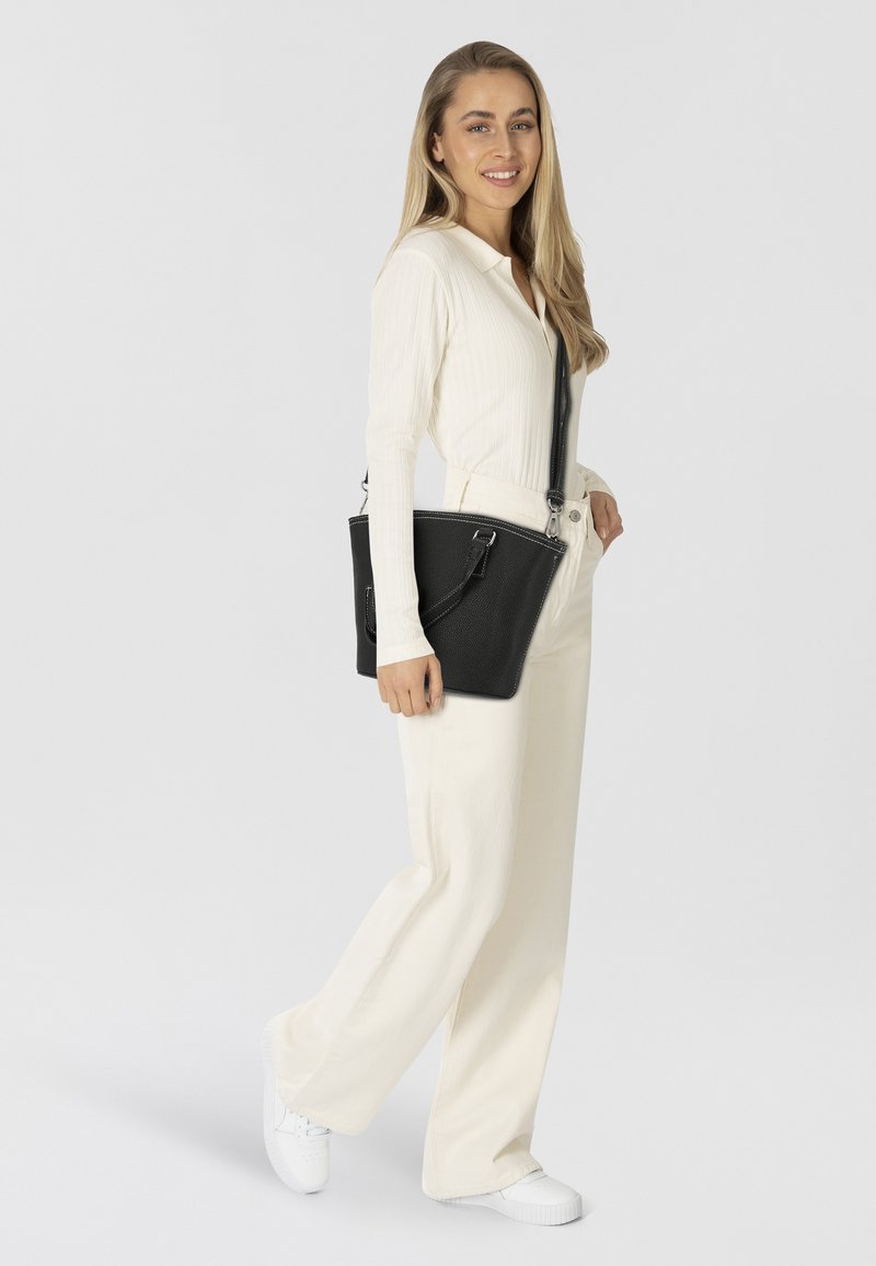 Vrouw met lang, blond haar, gekleed in een crèmekleurige wijde broek en trui, met witte sneakers, terwijl ze een zwarte leren schoudertas draagt en glimlacht.