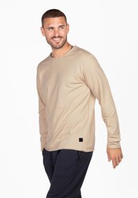 Beige Langarmshirt mit Rundhalsausschnitt und lockerer Passform. Mit einem kleinen schwarzen Logopatch am Saum und einem glatten, leichten Stoff.