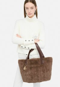 Borsa a tracolla in pelliccia ecologica marrone con manici in pelle marrone, texture a coste e dettagli in oro. La modella indossa un maglione bianco a coste e pantaloni bianchi.