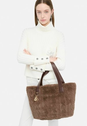 Bruine nep-bont handtas met bruine leren handvatten, geribbelde textuur en gouden hardware. Model draagt een witte geribbelde trui en witte broek.