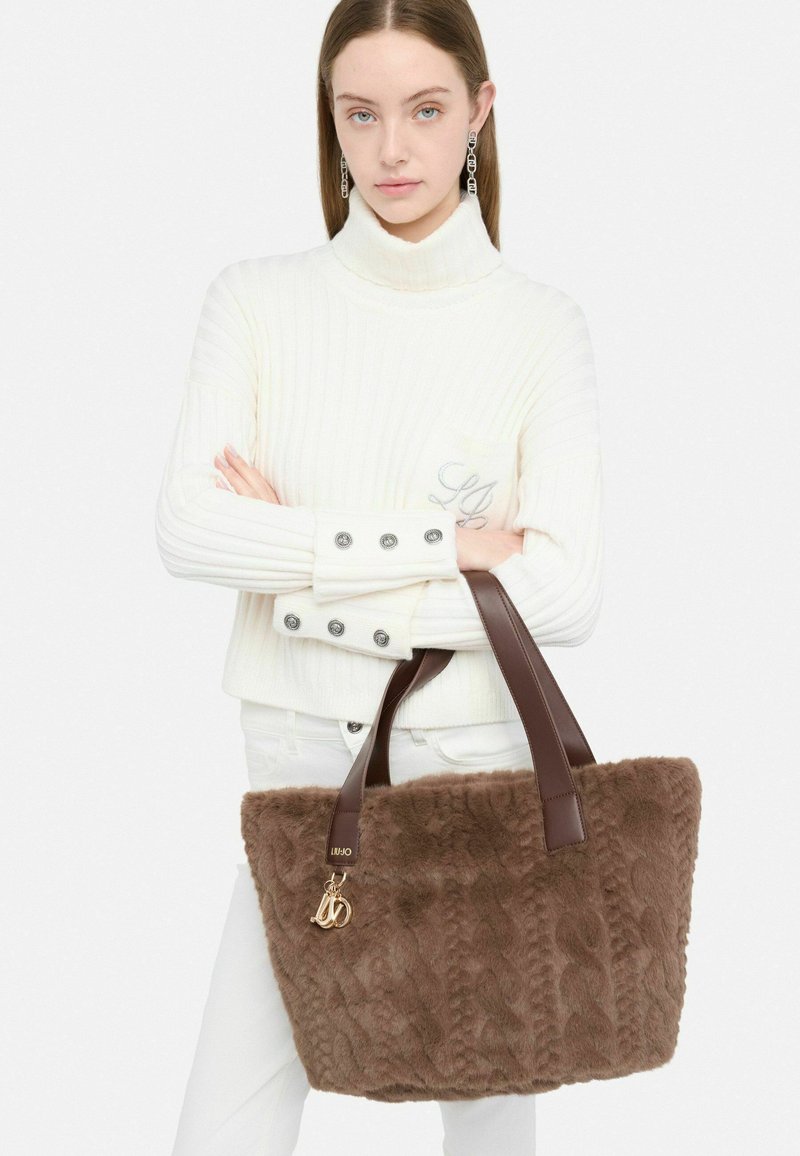 Borsa a tracolla in pelliccia ecologica marrone con manici in pelle marrone, texture a coste e dettagli in oro. La modella indossa un maglione bianco a coste e pantaloni bianchi.