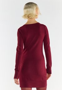 Blondynka z krótkimi włosami w długim rękawie, sukience do kolan w kolorze burgundowym, stojąca tyłem na jednolitym białym tle.