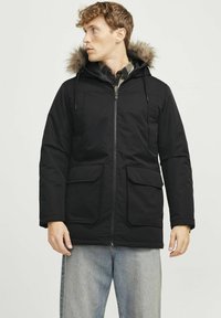 Sort parka med pelsforet hætte, lynlås foran, to store frontlommer og justerbare snore. Blødt, polstret stof med en mat finish.