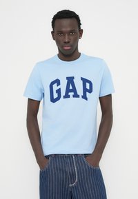 T-shirt en coton bleu clair avec logo "GAP" marine en lettres épaisses, associé à un short en denim rayé bleu foncé. Manches courtes et col rond.