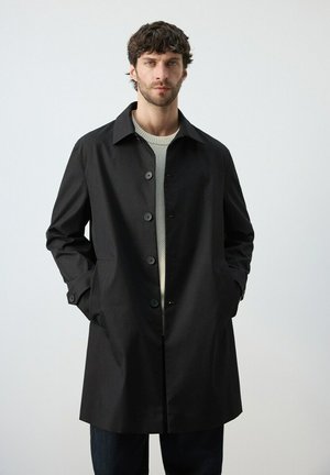 Mann trägt einen schwarzen, geknöpften Mantel über einem hellgrauen Pullover und steht mit den Händen in den Taschen vor einem schlichten hellen Hintergrund.