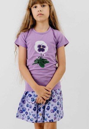 Young girl with long hair wearing a purple t-shirt with a large pansy flower and a matching floral skirt, standing with hands clasped.