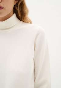 Maglione a collo alto color crema realizzato in un morbido tessuto a maglia con un design aderente e una texture liscia, caratterizzato da maniche lunghe e un alto colletto.