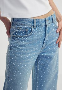 Vaqueros de mezclilla azul claro con embellishments de lunares blancos, corte recto, cinco bolsillos y cierre de botones.