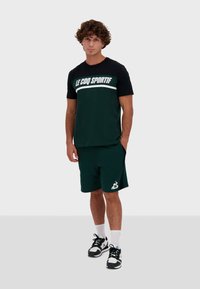 Camiseta deportiva verde con mangas negras y logo, combinada con pantalones cortos verdes a juego. Zapatillas negras y blancas, calcetines blancos.