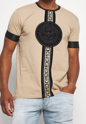 Beige kortærmet skjorte med sorte armbind, lodret sort-og-guld geometrisk stribe og rundt sort emblem med teksten "Glorious Gangsta".