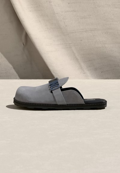 Γκρι mule slip-on με μαύρη σόλα και λουράκι με ασημένια μεταλλικά γράμματα στο πάνω μέρος, τοποθετημένο σε μπεζ υφασμάτινο φόντο.