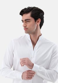 Camicia bianca a maniche lunghe con colletto alla coreana, caratterizzata da una texture liscia e una chiusura con bottoni. Il tessuto appare leggero e formale.