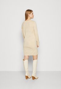 Šviesiai rudas megztas suknelė su ilgomis rankovėmis, laisvu kirpimu ir apvaliu iškirpimu, derinama su aukštais blizgiais šviesiai rudais batais su blokuoto kulno.